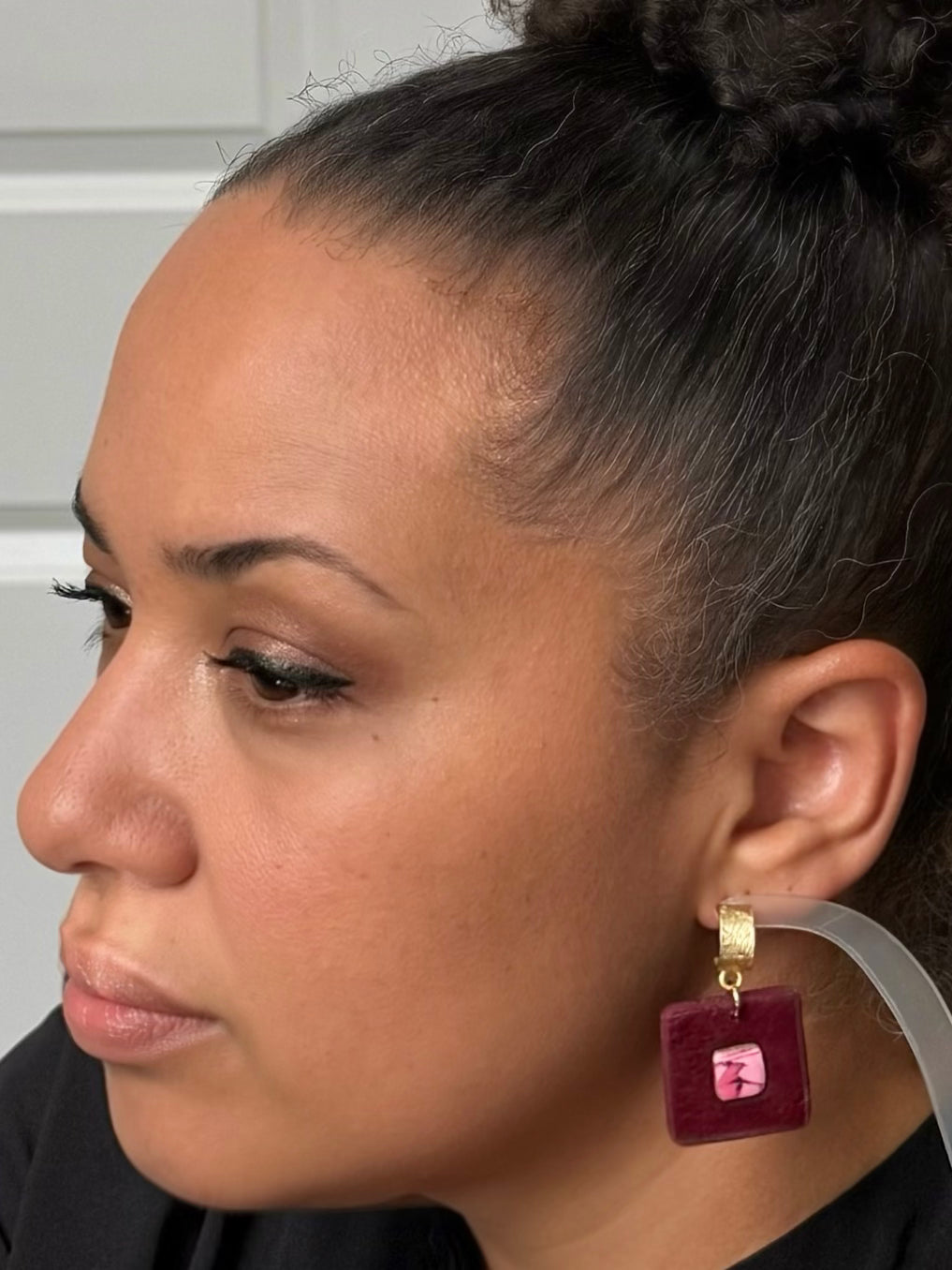 A close-up side profile of a woman wearing a dangling earring. The earring features a square burgundy velvet-like charm with a pink and black faux stone and gold-tone metal hardware.