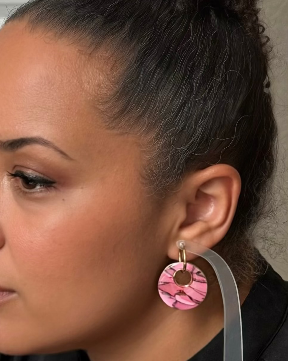 A close-up side profile of a woman wearing a pink and black patterned circular drop earring with a gold hoop
