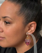 A close-up side profile of a woman wearing a dangling heart-shaped earring. The earring has a gold-tone top loop and a faceted purple-pink heart charm