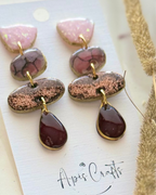A pair of handmade dangle earrings featuring a pink textured stone, a marbled purple and brown stone, a glittery rose gold stone, and a deep red teardrop drop.