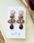 A pair of handmade dangle earrings featuring a pink textured stone, a marbled purple and brown stone, a glittery rose gold stone, and a deep red teardrop drop.