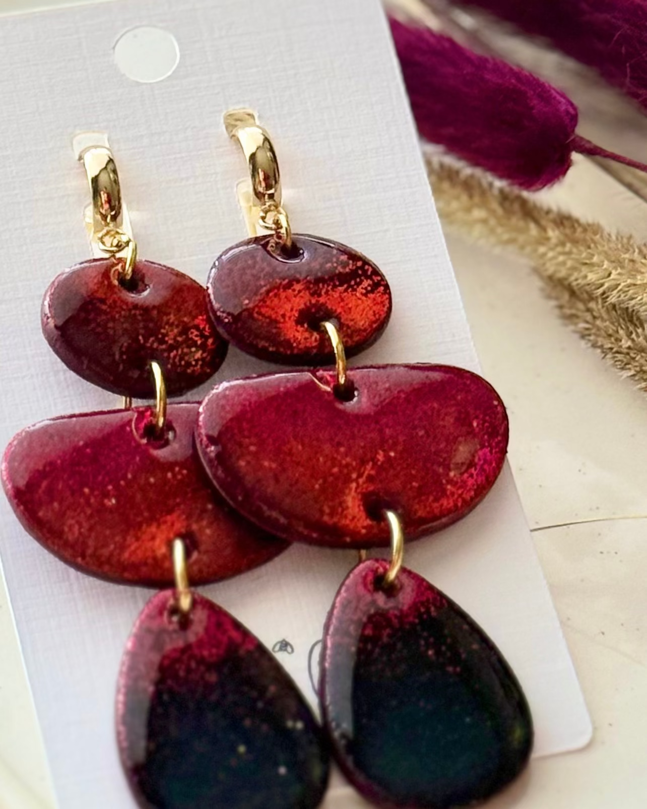 A pair of oversized dangling earrings with three stacked organic-shaped drops in a metallic red gradient with small gold clasps at the top.