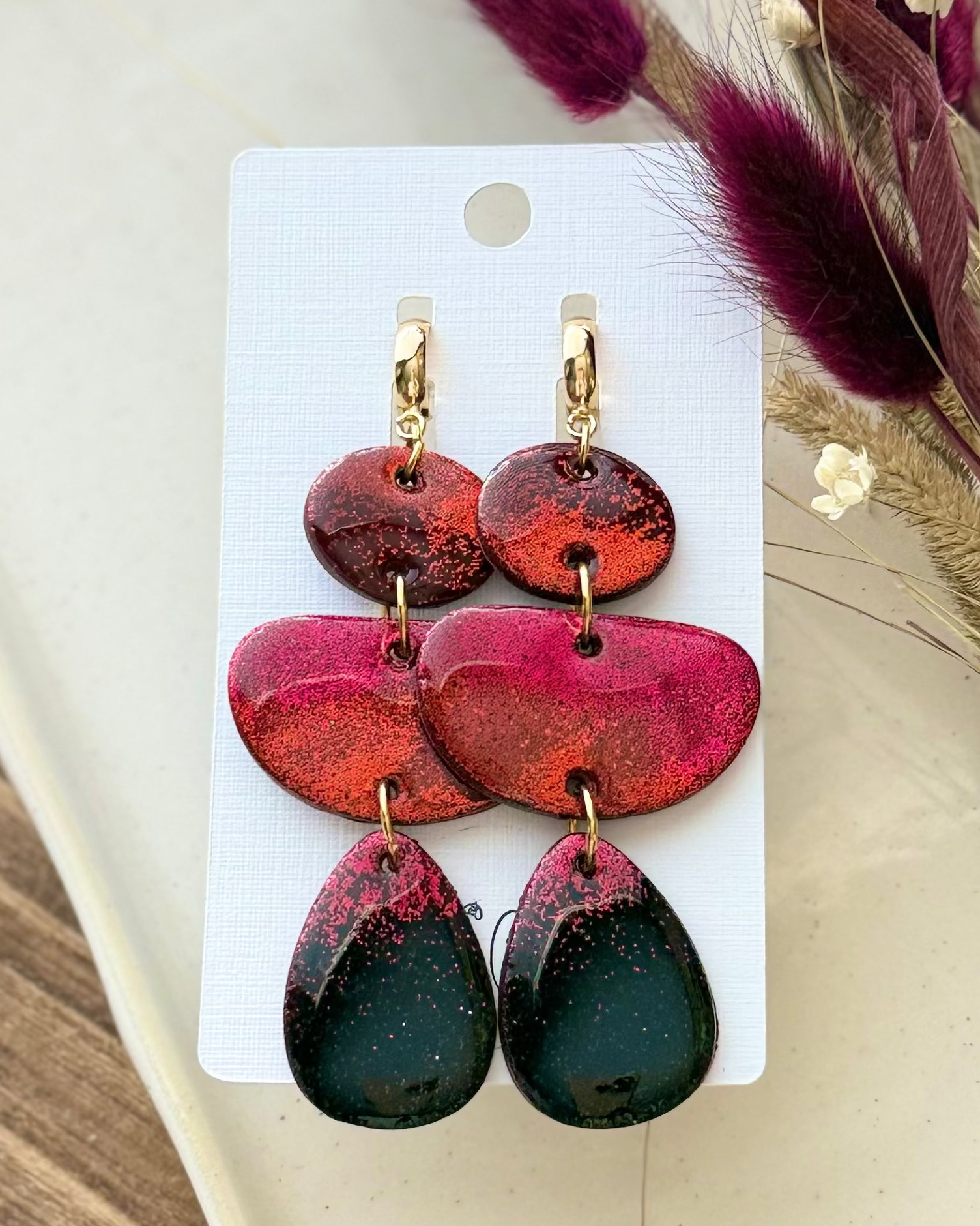 A pair of handmade dangle earrings with three organic shapes in a red ombré glitter design and small gold hoop closure