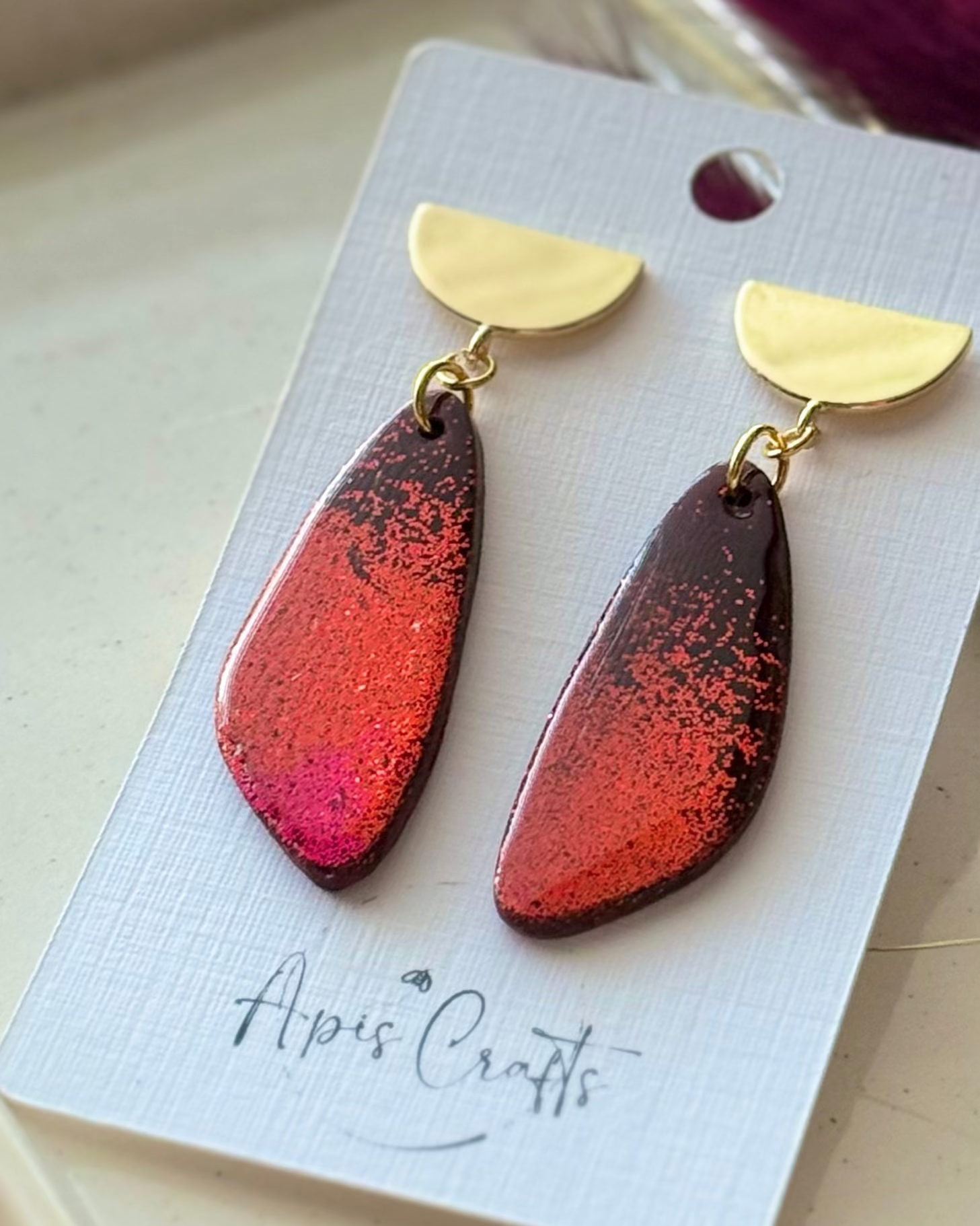 A pair of teardrop-shaped drop earrings with gold semicircle studs and a red ombré glittery gradient body