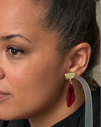 A close-up of a woman wearing a modern statement earring with a gold-toned semi-circle stud, a gold connector ring, and a teardrop-shaped deep red glitter drop