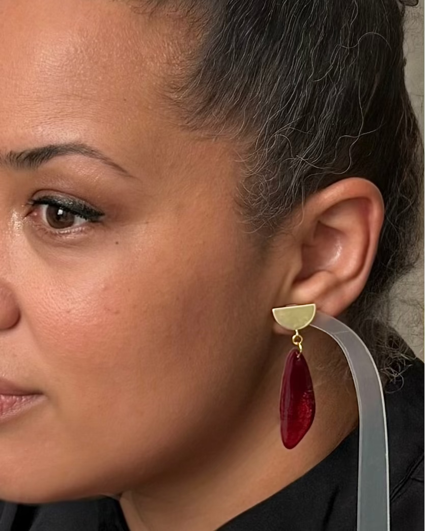 A close-up of a woman wearing a modern statement earring with a gold-toned semi-circle stud, a gold connector ring, and a teardrop-shaped deep red glitter drop