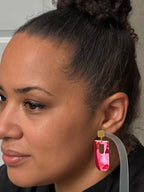 A close-up side profile of a woman wearing a chunky, geometric pink acrylic drop earring with a gold-tone connector