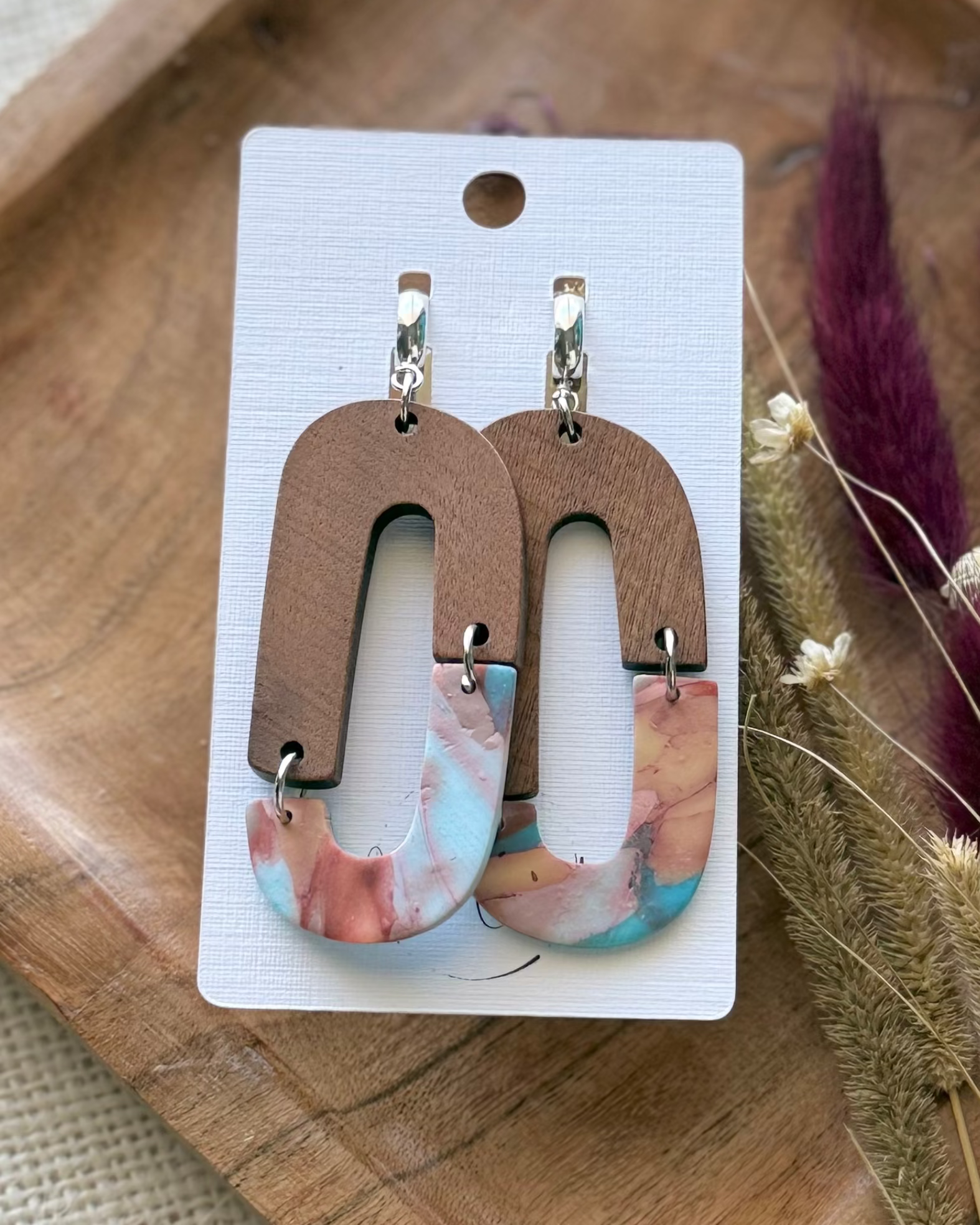A pair of statement earrings feature a brown wooden arch top and a dangling marbled resin bottom in pink, blue, and white.