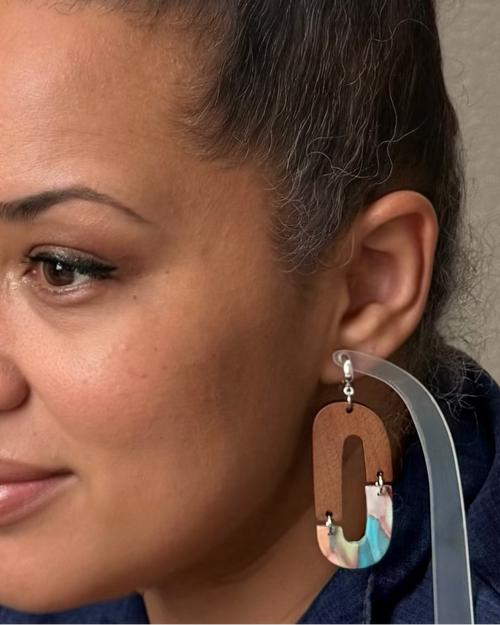 A close-up of a woman wearing a large, asymmetrical statement earring. The earring features a prominent brown wooden 'O' shape with a colorful marbled acrylic bottom section and a silver-toned hook.