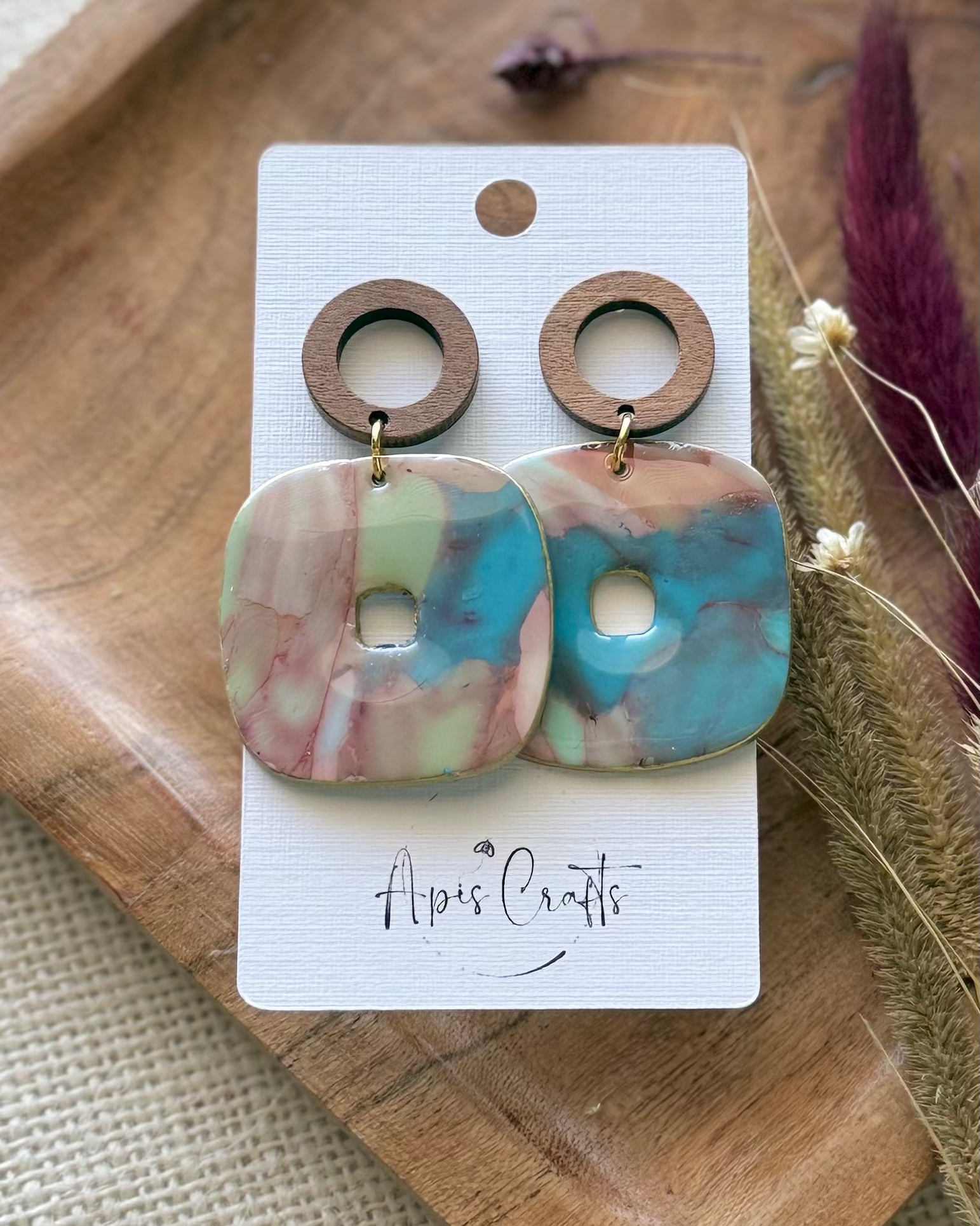 A pair of drop earrings with a round wooden hoop and a marbled square pendant in pink, turquoise blue, and pale green.