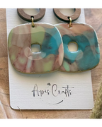 A pair of drop earrings with a round wooden hoop and a marbled square pendant in pink, turquoise blue, and pale green.