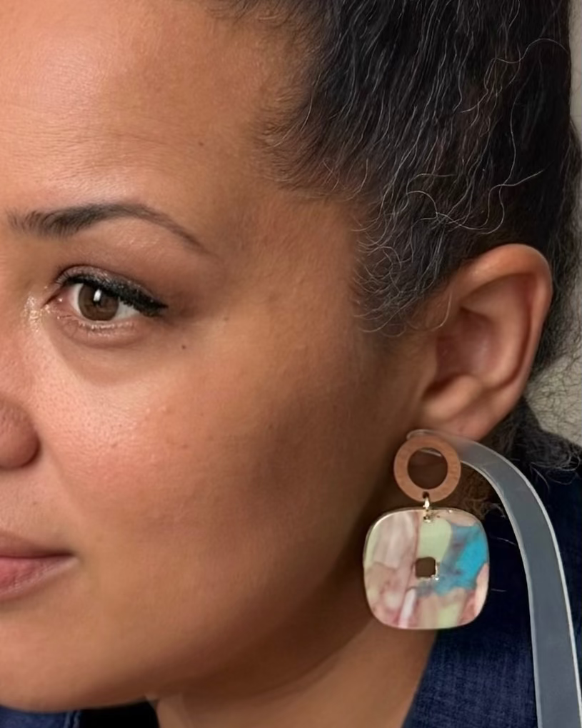 A close-up of a woman wearing a dangling earring with a round copper-toned top and a square marbled resin drop in pastel pink, blue, and cream.