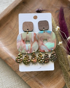 A pair of dangle featuring square marbled  pendants in pink, green, and white, with textured hammered teardrop charms in a bronze/gold finish, and wooden square studs.