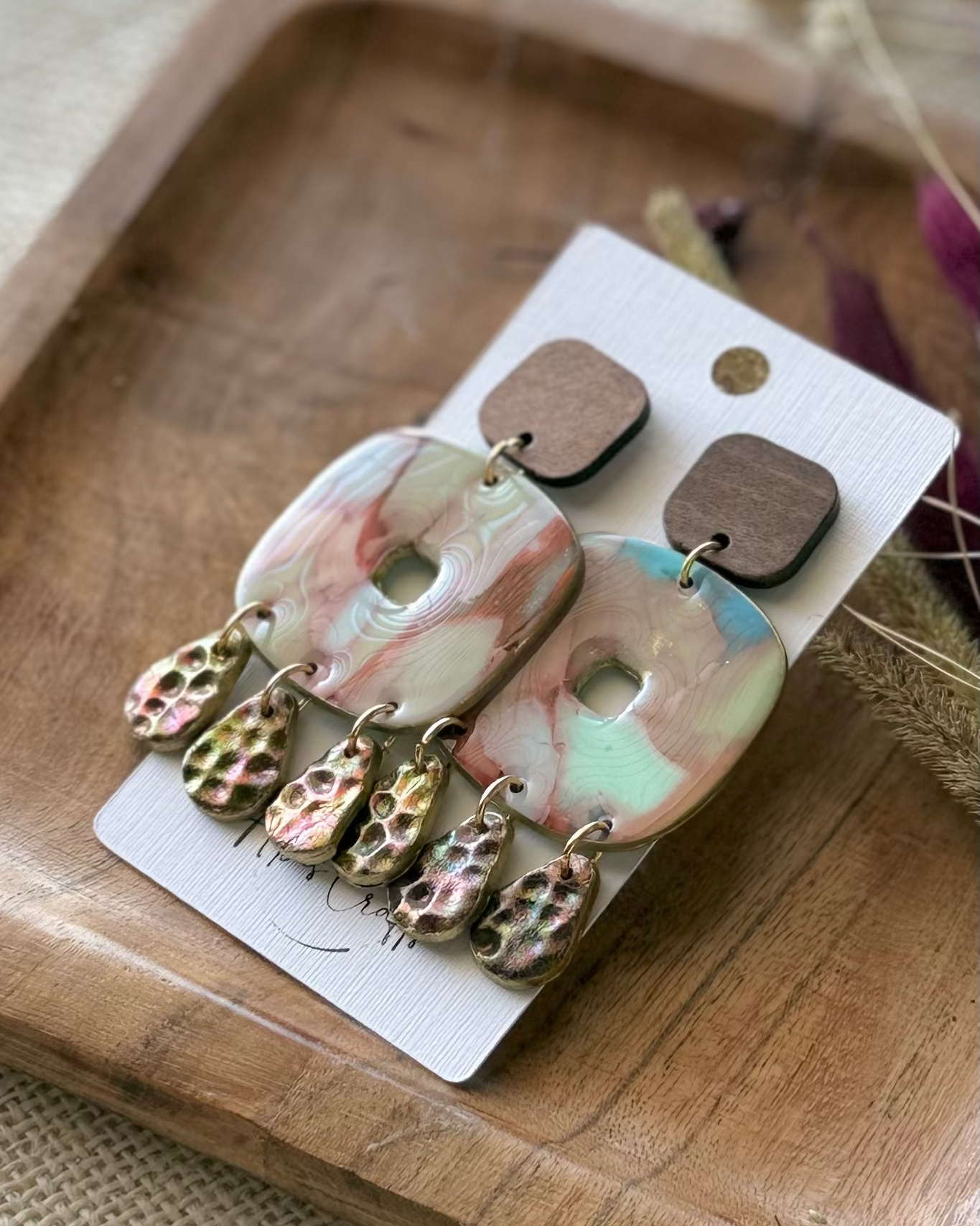 A pair of dangle featuring square marbled  pendants in pink, green, and white, with textured hammered teardrop charms in a bronze/gold finish, and wooden square studs.