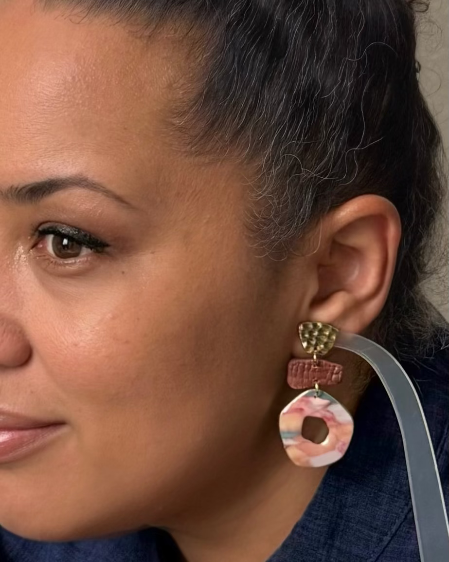 A close-up of a woman wearing a pair of chunky, multi-layered statement earrings. The earrings feature a textured gold-toned top, a reddish-brown middle section, and a circular bottom charm with pink, white, and orange marbled inclusions.