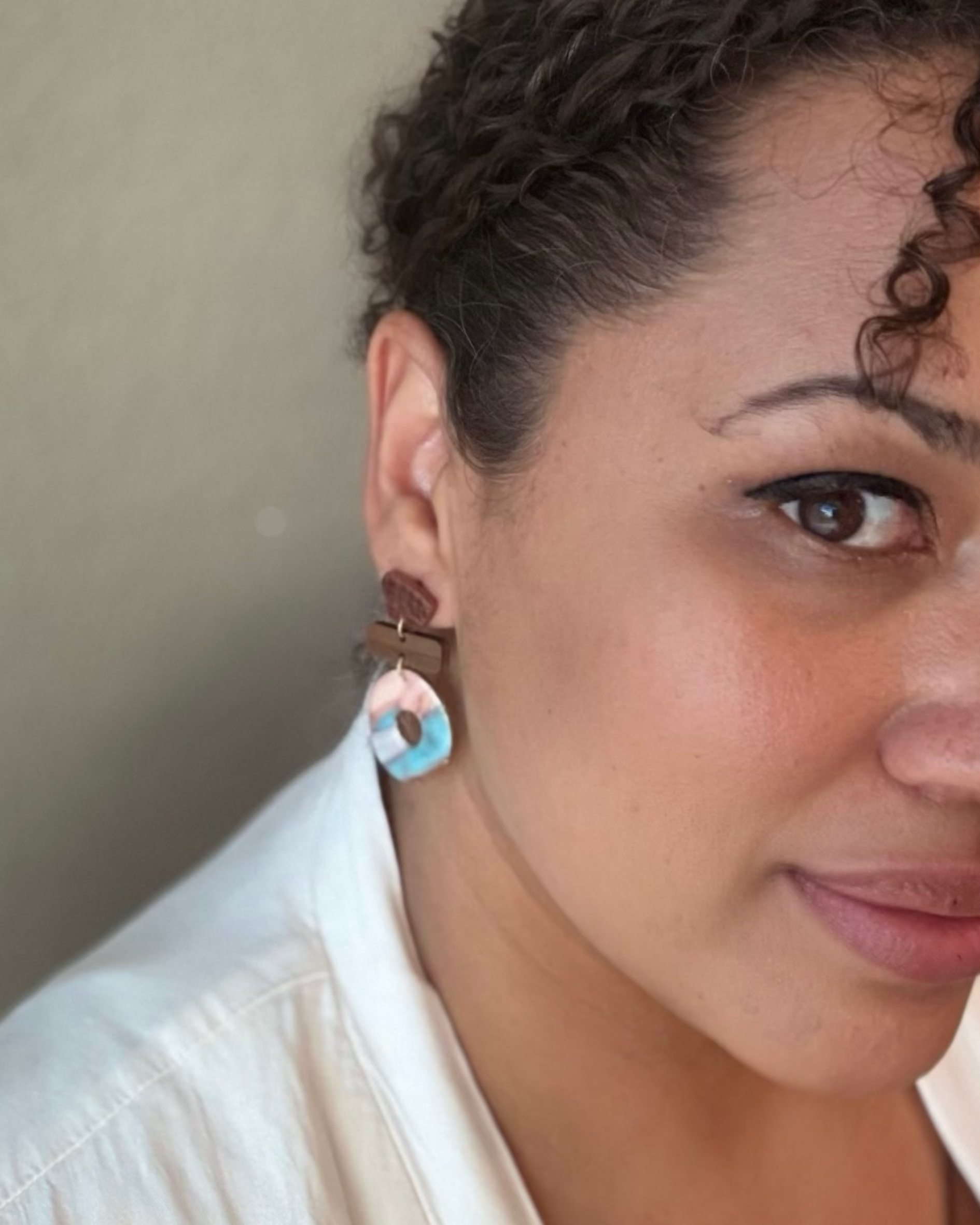 A close-up portrait of a woman wearing a dangling earring with a brown top and a circular charm featuring pastel blue, pink, and white concentric rings.