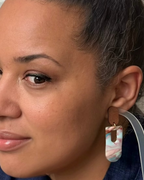 A close-up of a woman wearing a dangling earring. The earring features a brown leather top tab and a marbled acrylic drop in pastel pink, blue, and white, suspended from a gold-tone ring.