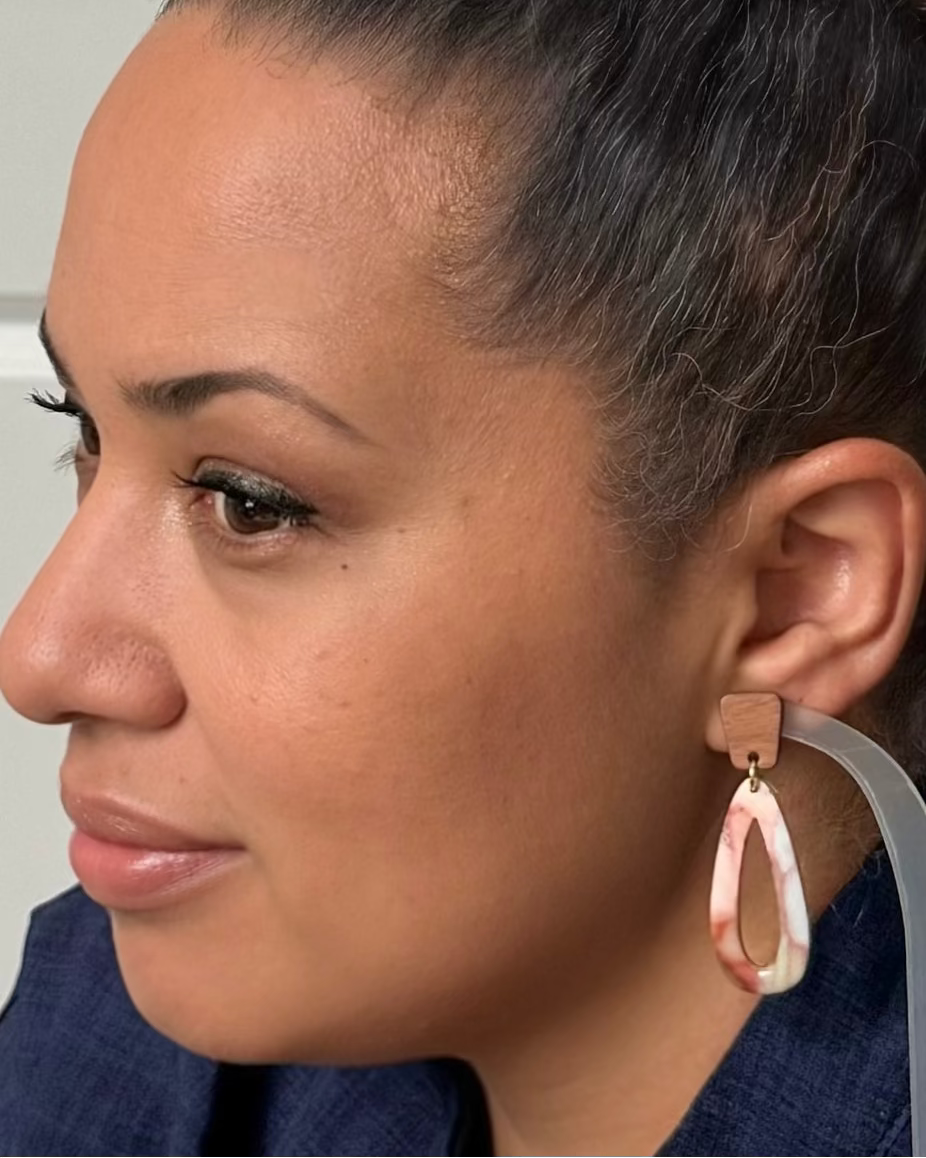 Close-up side profile of a woman wearing a teardrop-shaped dangling earring with a pink and white marbled pattern and a tan stud above it.