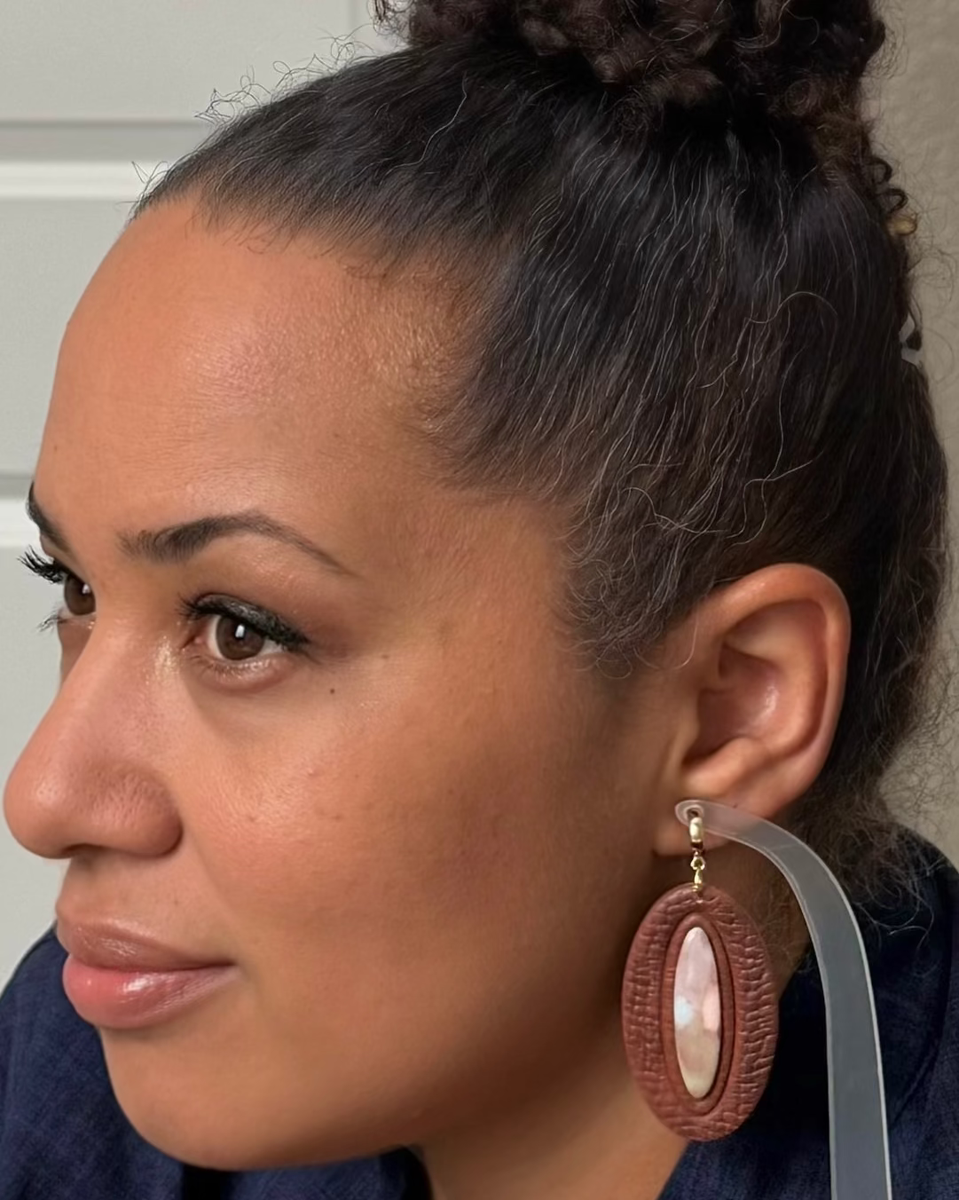 A close-up side profile of a woman wearing a large oval dangle earring with a brown woven-texture frame and a white mother-of-pearl center, shown against a neutral indoor background.