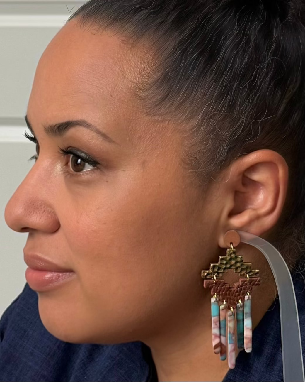 A close-up side profile of a woman wearing a large, intricate dangling earring. The earring features a bronze-toned geometric top, a brown woven band, and multiple dangling strands in turquoise blue, pink, and white marbled resin.