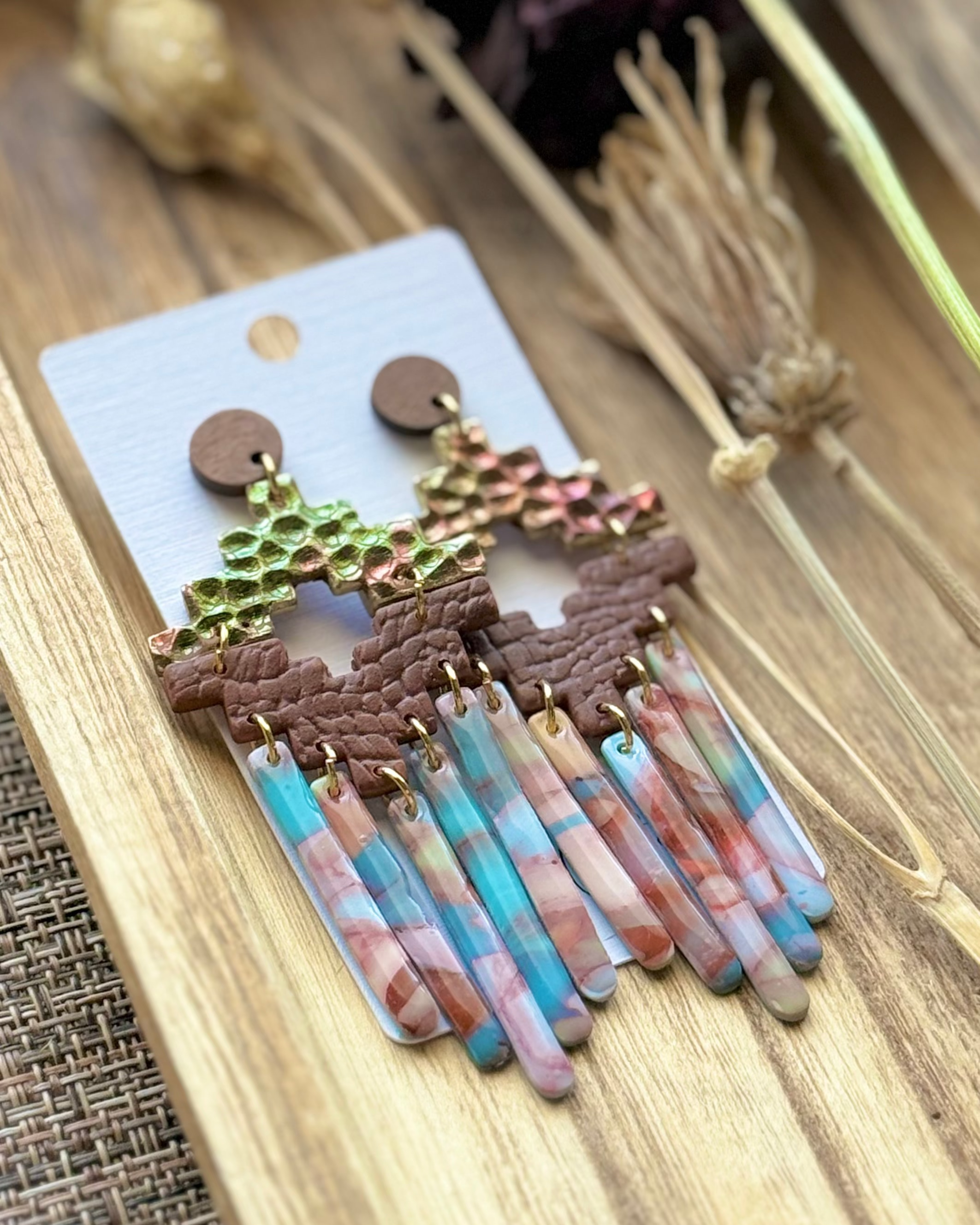 A pair of handmade statement earrings featuring textured gold and green top pieces, brown geometric middle bands, and dangling marbled fringe in pink, blue, and white.