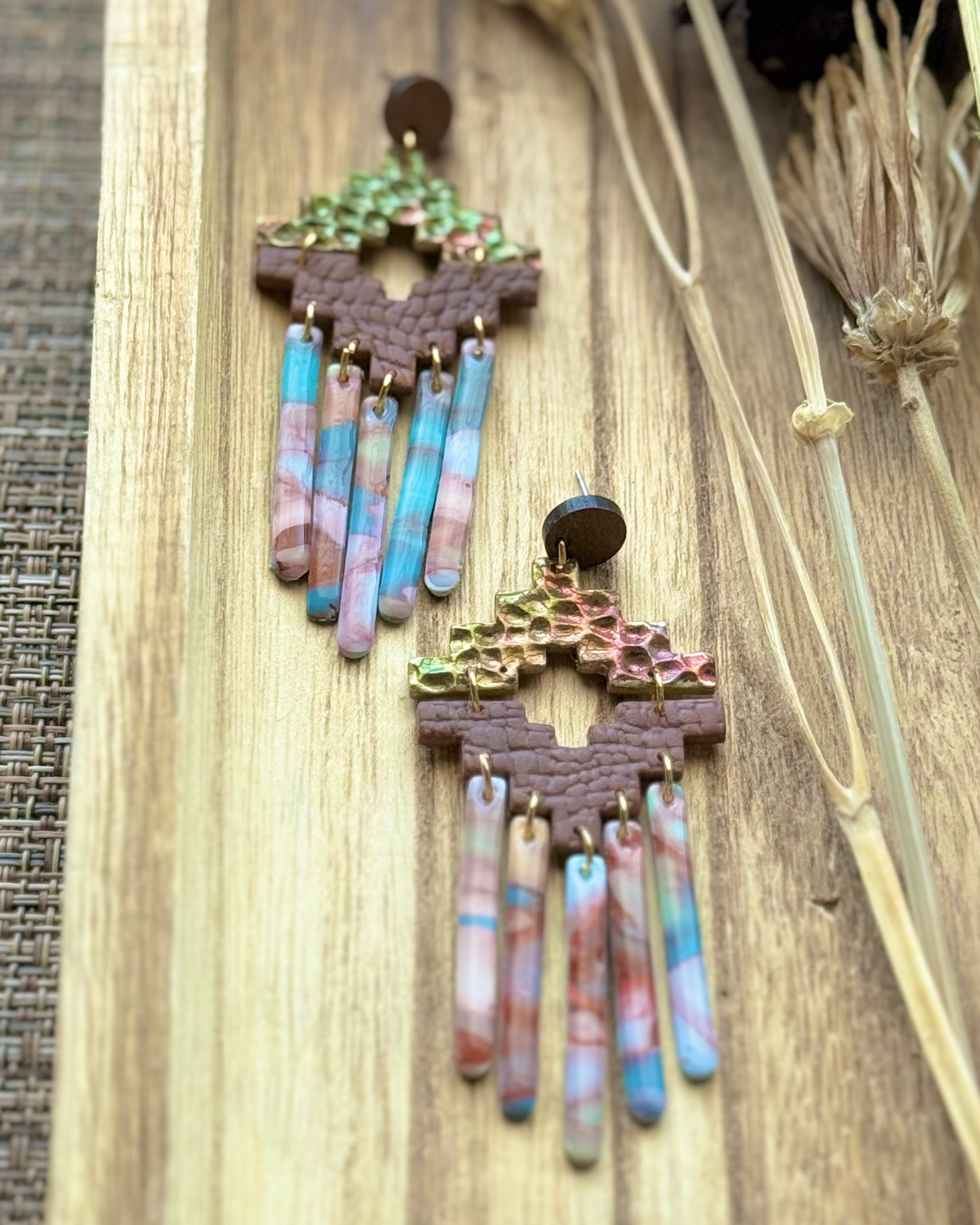 A pair of handmade statement earrings featuring textured gold and green top pieces, brown geometric middle bands, and dangling marbled fringe in pink, blue, and white.