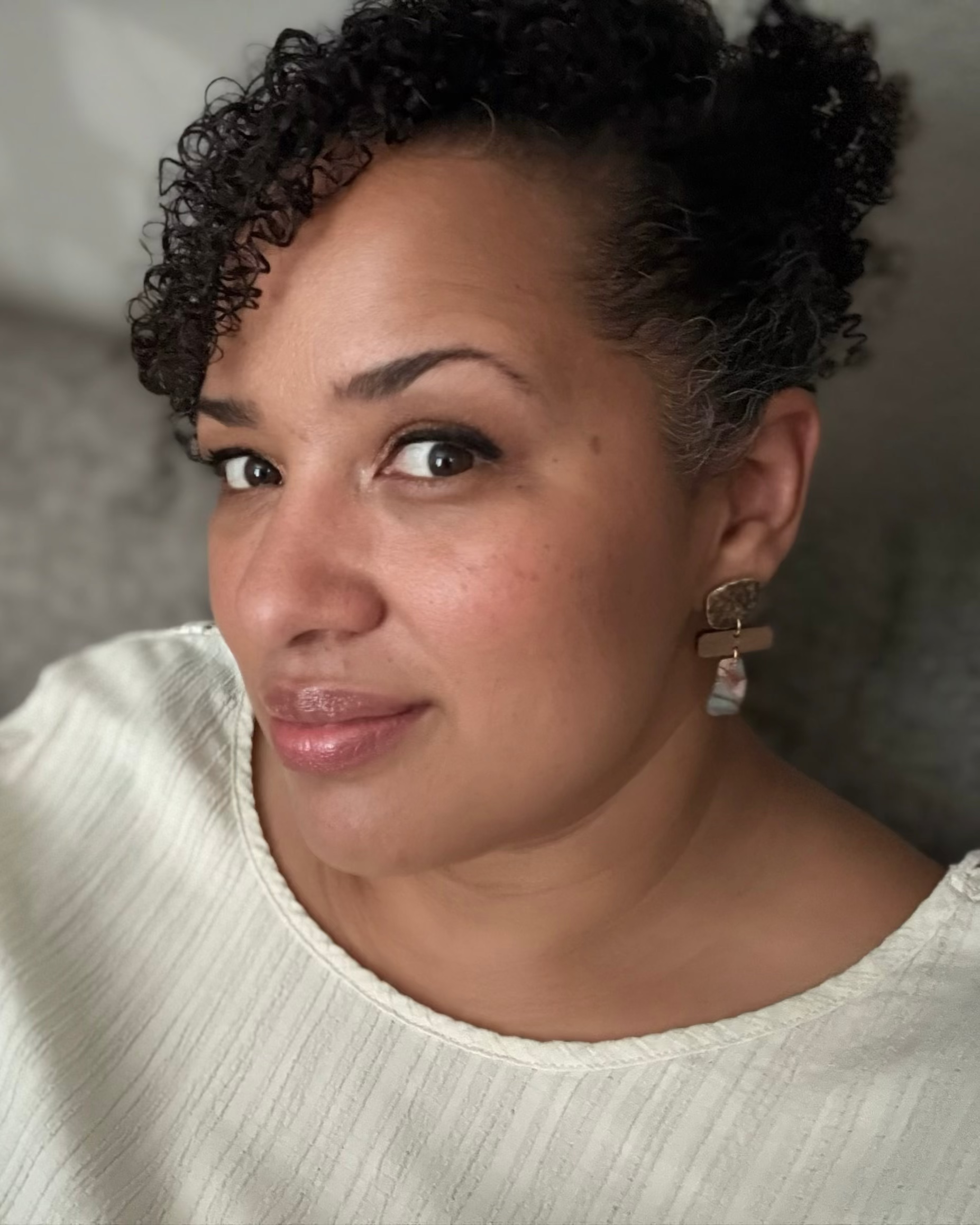 A close-up portrait of a woman with curly dark hair wearing a cream-colored ribbed top and a gold cross earring, seated against a gray upholstered background.