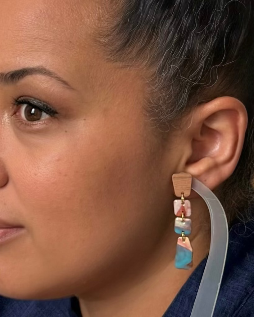 A close-up side profile of a woman wearing a long drop earring. The earring features a light brown wooden top, a pink and white marbled middle section, and a blue marbled bottom section, hanging from a clear curved band.