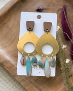 A pair of statement dangle earrings featuring a large gold-toned circular disc with a cutout, a small brown wooden top piece, and three dangling marbled resin teardrop charms in pink, blue, and white.