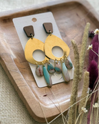 A pair of statement dangle earrings featuring a large gold-toned circular disc with a cutout, a small brown wooden top piece, and three dangling marbled resin teardrop charms in pink, blue, and white.