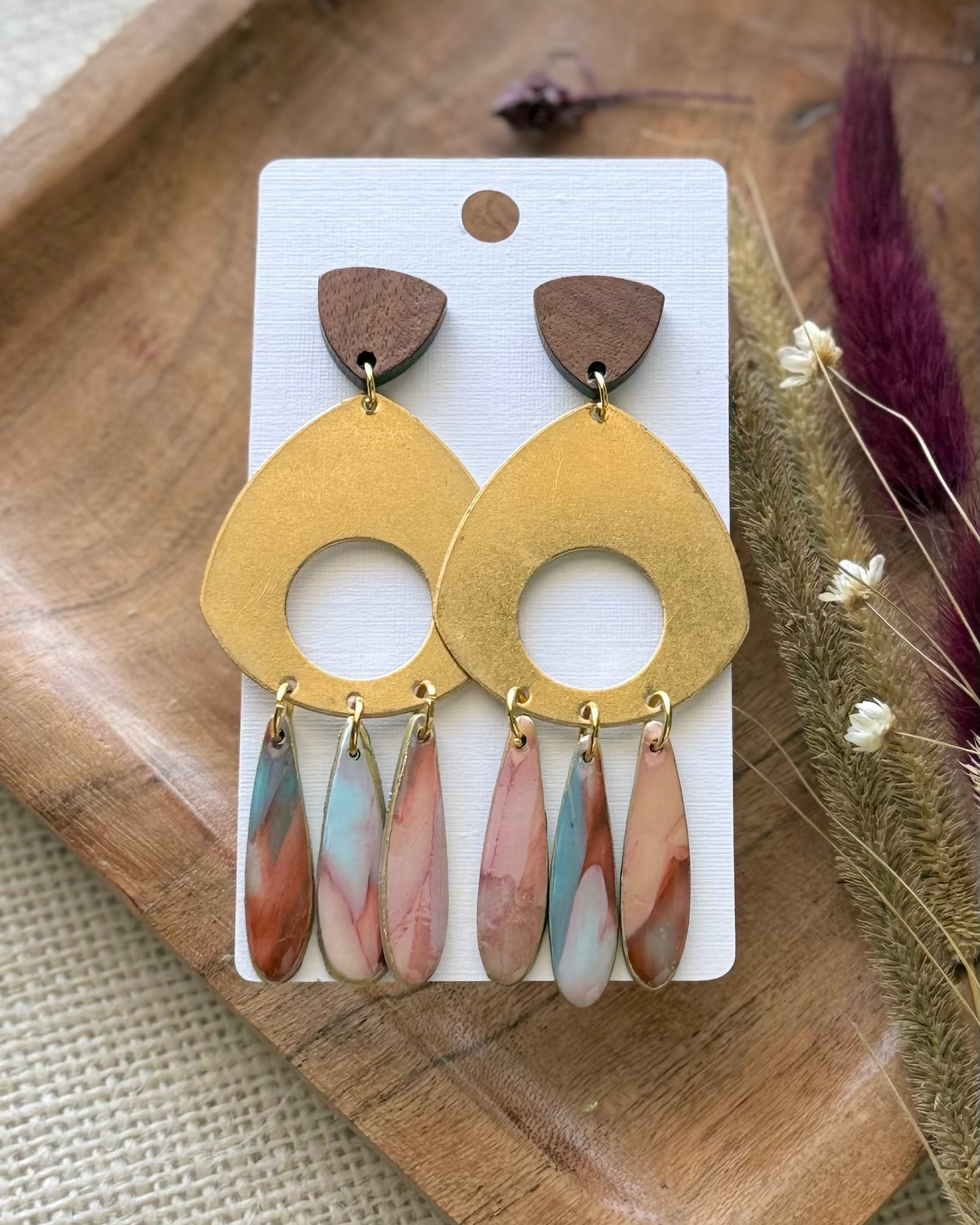 A pair of statement dangle earrings featuring a large gold-toned circular disc with a cutout, a small brown wooden top piece, and three dangling marbled resin teardrop charms in pink, blue, and white.