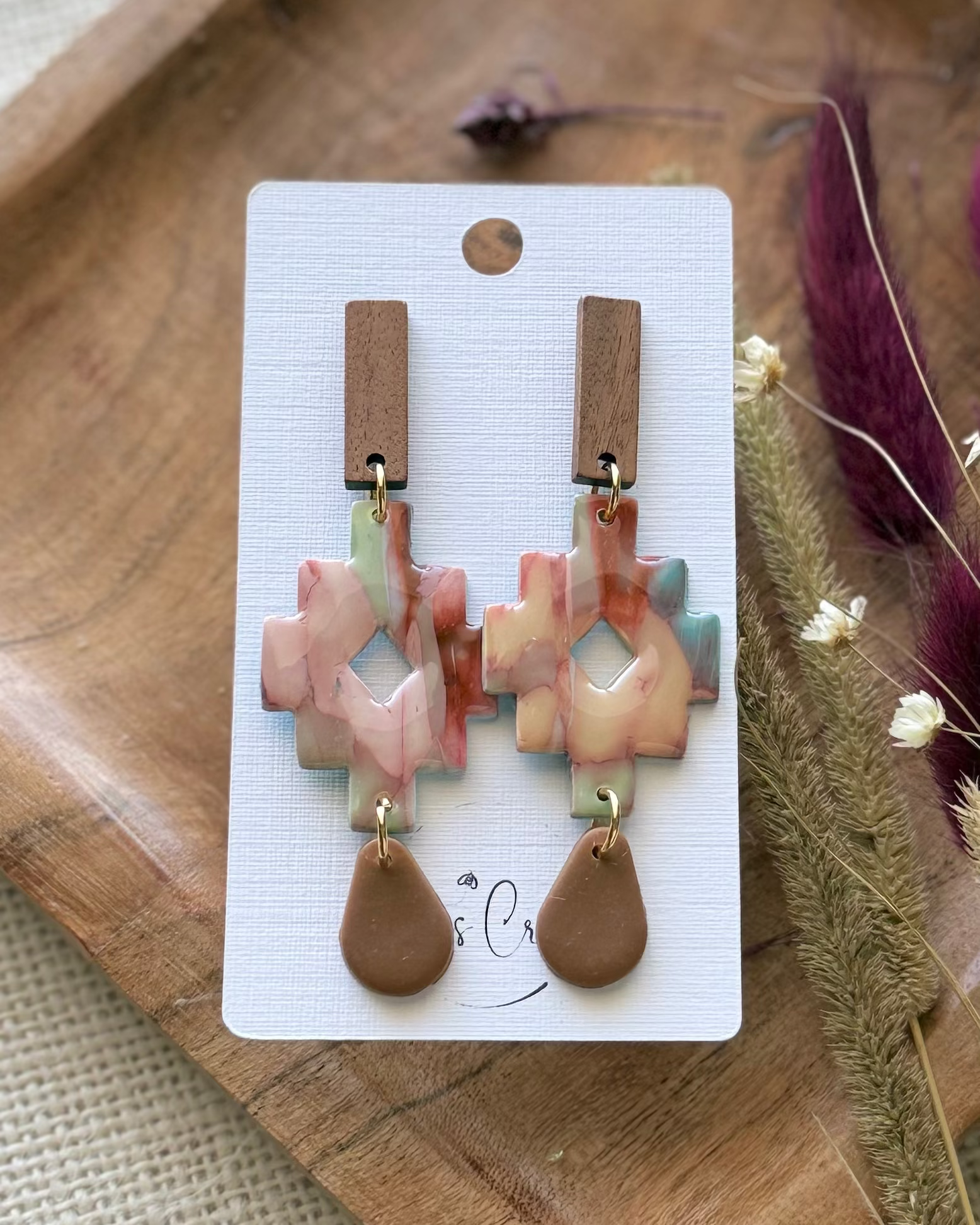 A pair of handmade dangle earrings featuring a geometric cross-shaped charm with a marbled pink, beige, and light blue pattern, and a brown teardrop drop.