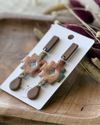 A pair of handmade dangle earrings featuring a geometric cross-shaped charm with a marbled pink, beige, and light blue pattern, and a brown teardrop drop.