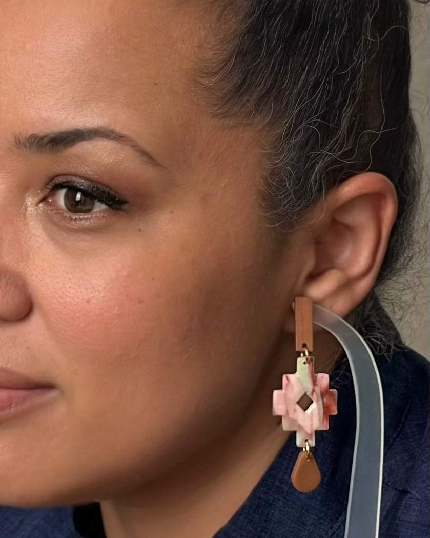 A close-up of a woman wearing a dangling geometric earring with pink, white, and brown elements, shown against her ear and dark hair.