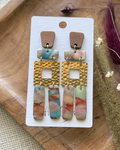 A pair of bohemian-style drop earrings featuring a brown wood-grain top, gold-tone hammered square links, and marbled drops in pink, blue, and green.