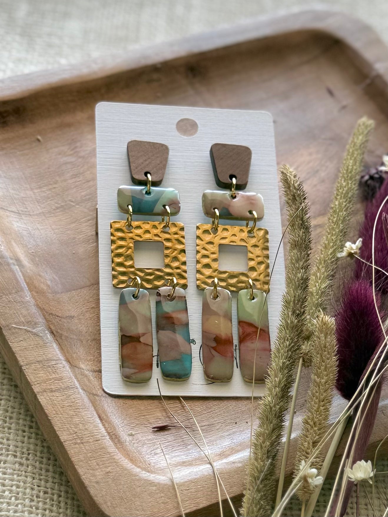A pair of bohemian-style drop earrings featuring a brown wood-grain top, gold-tone hammered square links, and marbled drops in pink, blue, and green.