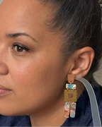 A close-up side profile of a woman wearing a long, multi-tiered statement earring. The earring features a tan leather top, a gold-toned geometric frame, and dangling turquoise and marbled blue, white, and pink beads.