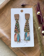 A pair of handmade dangle earrings feature a textured, hammered metallic top section with iridescent rainbow reflections, a small brown wooden square, and two dangling fan-shaped pieces in pink and blue.