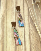 A pair of handmade dangle earrings feature a textured, hammered metallic top section with iridescent rainbow reflections, a small brown wooden square, and two dangling fan-shaped pieces in pink and blue.