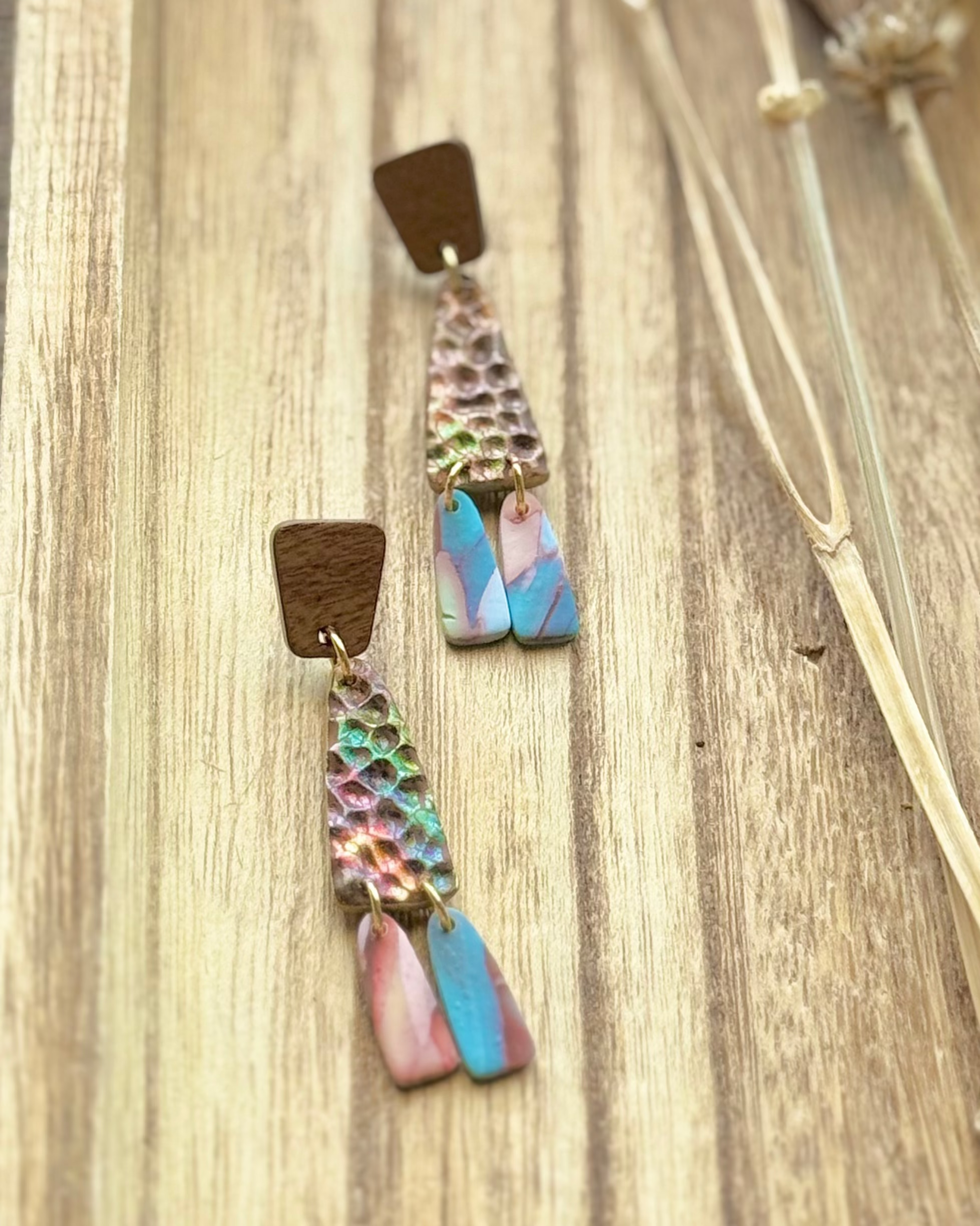 A pair of handmade dangle earrings feature a textured, hammered metallic top section with iridescent rainbow reflections, a small brown wooden square, and two dangling fan-shaped pieces in pink and blue.