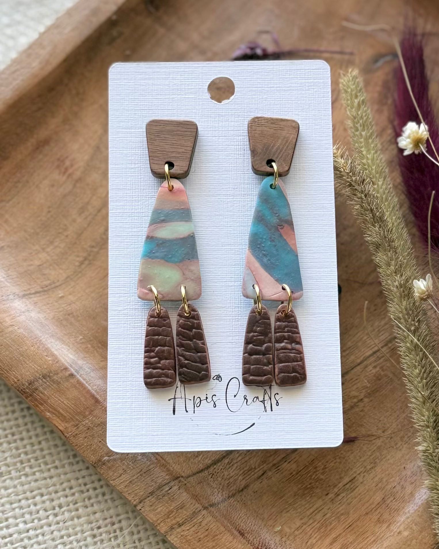 A pair of handmade dangle earrings feature a textured, hammered metallic top section with iridescent rainbow reflections, a small brown wooden square, and two dangling fan-shaped pieces in pink and blue.