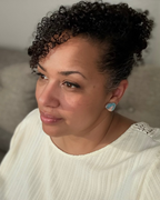 A close-up portrait of a woman with short, curly dark hair wearing a matching curly hair extension or wig, a white ribbed top, and a light blue heart-shaped earring, set against a soft-focus gray indoor background.