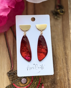 A pair of dangle earrings displayed on a white card. Each earring has a gold semicircle stud and a dangling glittery red teardrop-shaped drop.