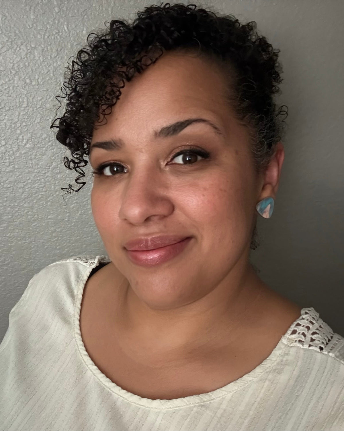 A woman with short curly dark hair wearing a white lace-trimmed top and a light blue heart-shaped earring, photographed against a textured gray wall.