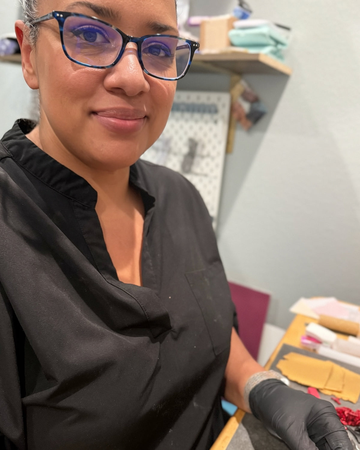 A woman with glasses and a black top wearing a black glove, holding a small item in a workshop or studio setting.
