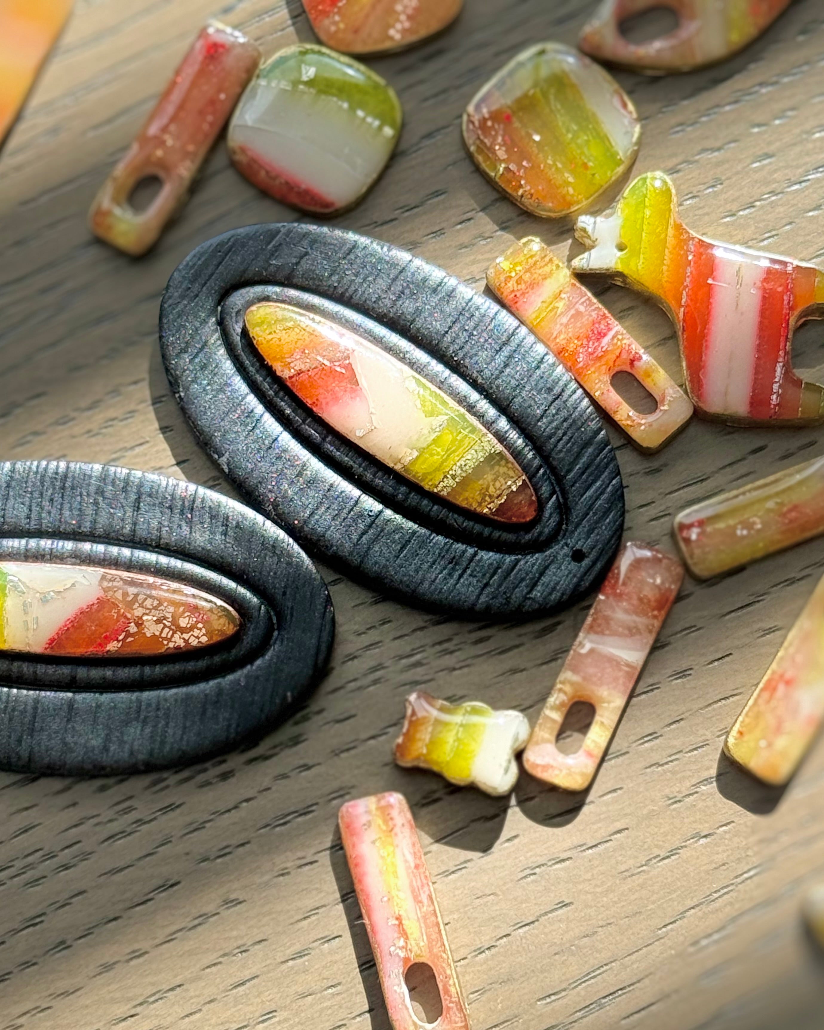 Two oval black earrings with colorful striped faux stone inserts, displayed on a wooden surface alongside several loose matching earrings.