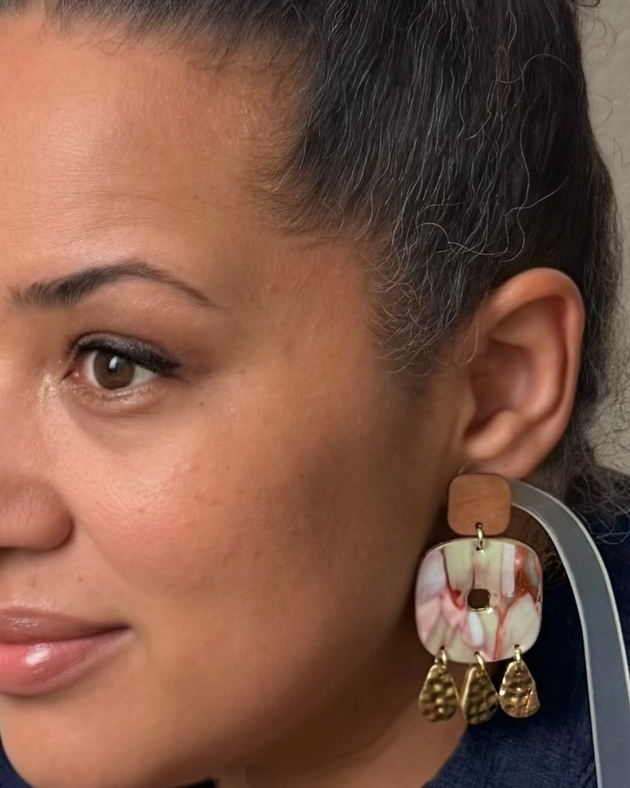 A close-up of a woman wearing a pair of large statement earrings. The earrings feature a square marbled shell-like charm in pink, white, and brown, with a tan leather top and dangling gold-toned textured charms.